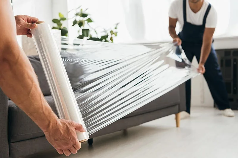 A worker covering furniture with plastic sheeting and taping edges to shield unaffected areas during restoration.